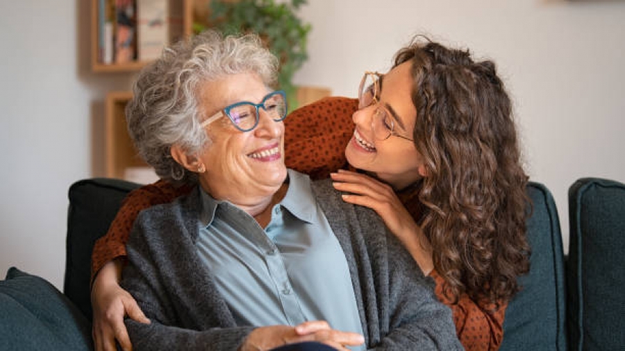 Old grandmother and adult granddaughter hugging at home and looking at each other. Happy senior mother and young daughter embracing with love on sofa. Happy young woman hugging from behind grandma with love.